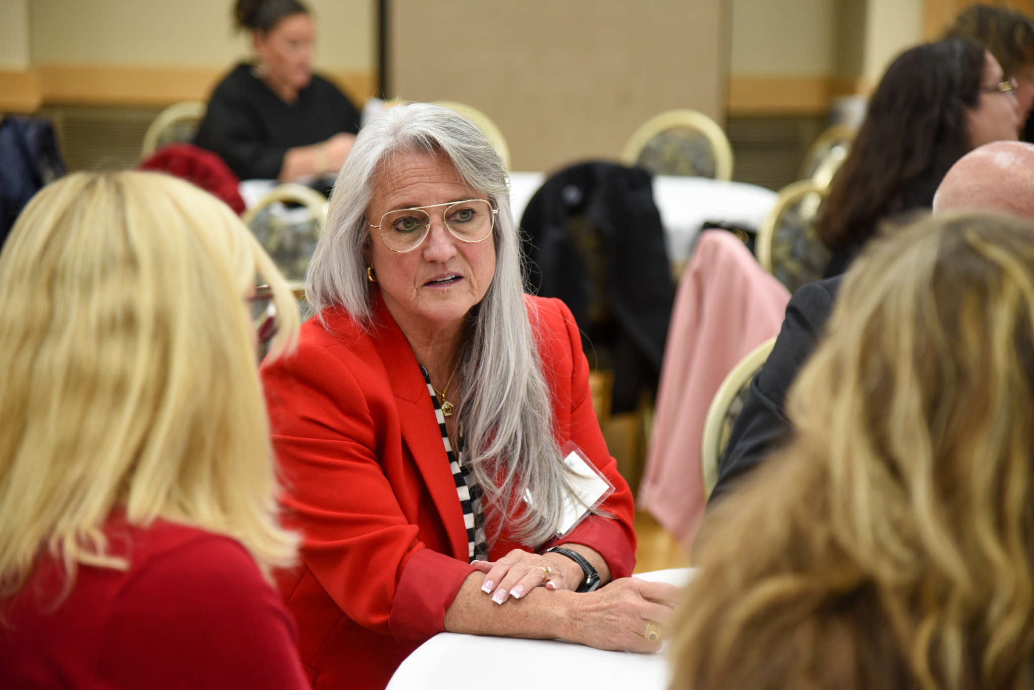women with long gray hair, glasses and a red blazer speaking with two other women at a table