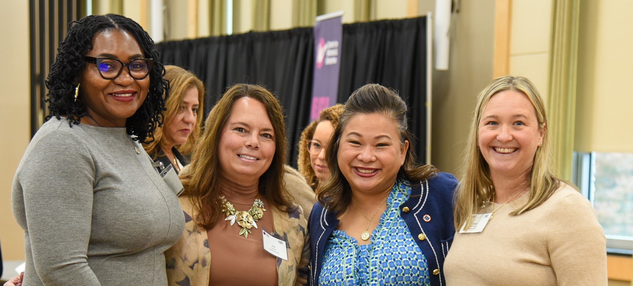 4 women standing together looking at camera with two women standing behind them talking